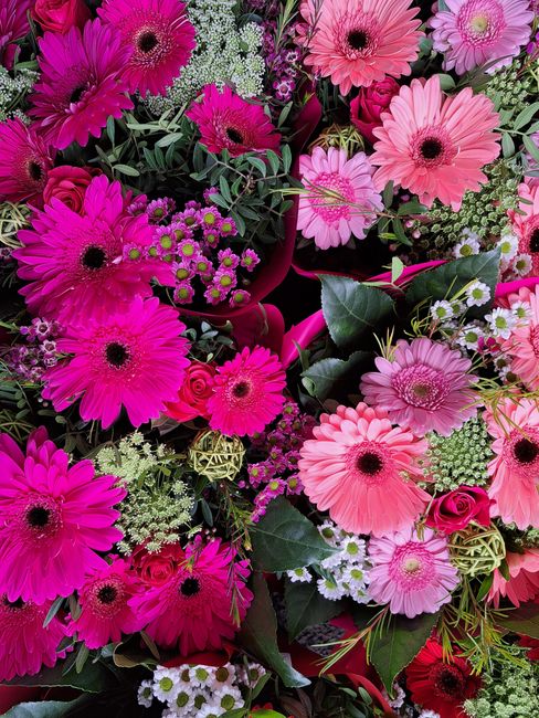 A vibrant floral arrangement featuring an assortment of pink and magenta gerbera daisies, complemented by small white and yellow filler flowers, dark green foliage, and wrapped in pink paper with a ma