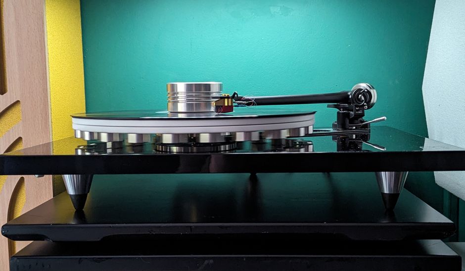 A close-up of a modern turntable with a black glossy finish, silver platter, and black tonearm resting on a black shelf. The turntable is set against a vibrant green wall with a textured surface, and 