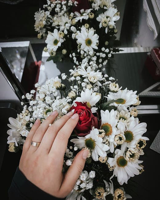 A floral arrangement featuring a mix of white daisies with yellow centers and delicate white baby's breath, arranged with a single deep red rose at the center. The flowers are held in a person's hand,