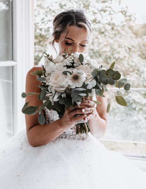 A bride holding a large floral bouquet with white roses, white daisies with dark centers, and eucalyptus leaves, wrapped in a satin ribbon at the base. She is seated indoors near a large window, with 